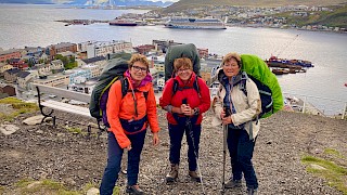 3 damer på en høyde og med utsikt over by og hav. Skyer på himmelen over. En cruisebåt på sjøen.